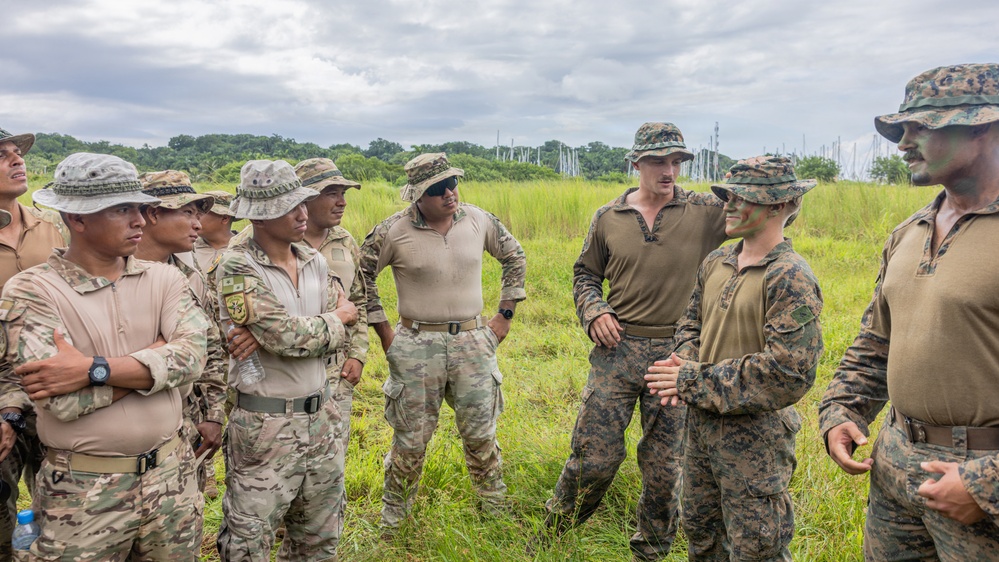 A group of military personnel in mixed camouflage uniforms are gathered in an open grassy field, appearing to be engaged in a briefing or discussion. Two distinct uniform types are visible: tan/desert camouflage worn by one group and woodland MARPAT digital camouflage worn by others, suggesting a joint multinational military exercise or training event. One individual wearing face camouflage paint appears to be addressing or conversing with the assembled group.