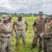 A group of military personnel in mixed camouflage uniforms are gathered in an open grassy field, appearing to be engaged in a briefing or discussion. Two distinct uniform types are visible: tan/desert camouflage worn by one group and woodland MARPAT digital camouflage worn by others, suggesting a joint multinational military exercise or training event. One individual wearing face camouflage paint appears to be addressing or conversing with the assembled group.