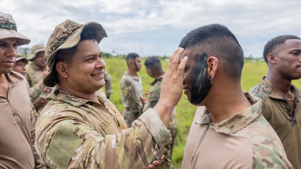 A smiling soldier in MultiCam/OCP camouflage uniform and boonie hat applies face camouflage paint to another soldier's face during what appears to be a field training exercise. Multiple military personnel in various camouflage uniforms are visible in the background on an open grassy field. The activity suggests pre-mission or training preparation involving camouflage application.