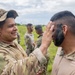 A smiling soldier in MultiCam/OCP camouflage uniform and boonie hat applies face camouflage paint to another soldier's face during what appears to be a field training exercise. Multiple military personnel in various camouflage uniforms are visible in the background on an open grassy field. The activity suggests pre-mission or training preparation involving camouflage application.