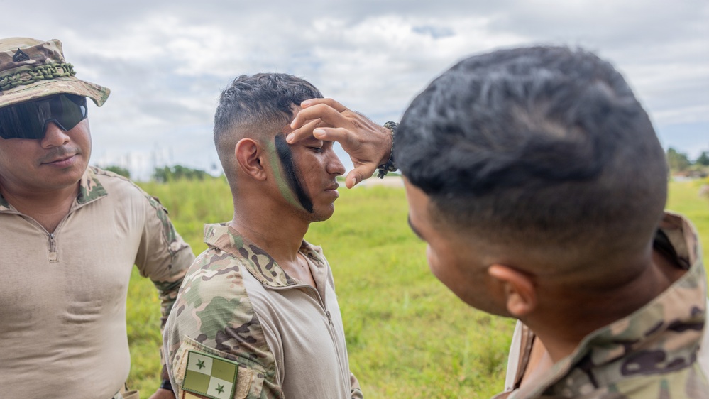 Military personnel are applying camouflage face paint to a soldier in an outdoor field setting. One soldier stands still while another applies green and black camouflage paint to his face, with a third soldier observing nearby. The setting appears to be a training exercise or pre-mission preparation.