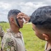 Military personnel are applying camouflage face paint to a soldier in an outdoor field setting. One soldier stands still while another applies green and black camouflage paint to his face, with a third soldier observing nearby. The setting appears to be a training exercise or pre-mission preparation.
