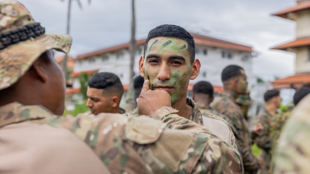 Military personnel in camouflage uniforms are applying face paint to one another outdoors, likely in preparation for a field exercise or training operation. The central subject is a young soldier having green and black camouflage face paint applied to his chin and cheek area by another service member. The setting appears to be a military installation with white buildings featuring red-tiled roofs visible in the background.