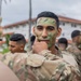 Military personnel in camouflage uniforms are applying face paint to one another outdoors, likely in preparation for a field exercise or training operation. The central subject is a young soldier having green and black camouflage face paint applied to his chin and cheek area by another service member. The setting appears to be a military installation with white buildings featuring red-tiled roofs visible in the background.