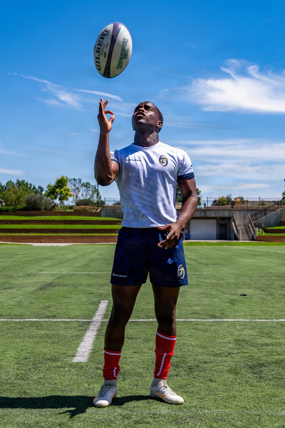 A rugby player wearing a white Yakuma jersey and navy blue shorts stands on a green turf field, tossing a Gilbert rugby ball into the air while looking upward. The athlete is dressed in full kit including red knee-high socks and white cleats. The photo is taken outdoors under a bright blue sky with light clouds.