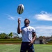 A rugby player wearing a white Yakuma jersey and navy blue shorts stands on a green turf field, tossing a Gilbert rugby ball into the air while looking upward. The athlete is dressed in full kit including red knee-high socks and white cleats. The photo is taken outdoors under a bright blue sky with light clouds.