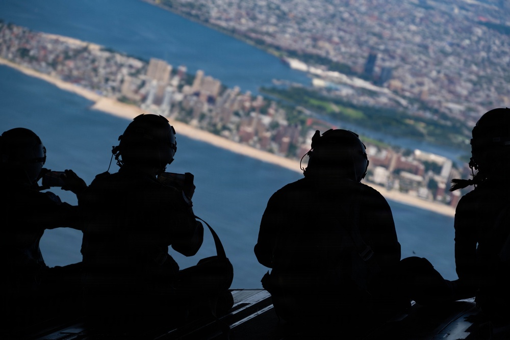 Four silhouetted individuals wearing flight helmets and communication headsets are seated inside an open military aircraft, looking out over a large coastal city from high altitude. The aerial view below shows a densely populated urban area with a prominent beach/shoreline, a river or bay, and what appears to be a large rectangular park. At least one person is holding a camera or device to document the view.