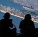 Four silhouetted individuals wearing flight helmets and communication headsets are seated inside an open military aircraft, looking out over a large coastal city from high altitude. The aerial view below shows a densely populated urban area with a prominent beach/shoreline, a river or bay, and what appears to be a large rectangular park. At least one person is holding a camera or device to document the view.