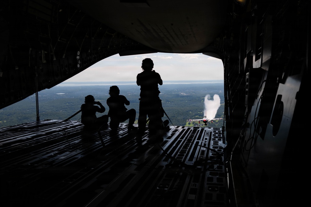 Three silhouetted individuals are seated and standing on the open rear cargo ramp of a large military transport aircraft in flight. They are observing or photographing another aircraft visible outside, which appears to be emitting a white smoke trail. The landscape below shows a forested area with what appears to be a body of water in the distance.