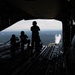 Three silhouetted individuals are seated and standing on the open rear cargo ramp of a large military transport aircraft in flight. They are observing or photographing another aircraft visible outside, which appears to be emitting a white smoke trail. The landscape below shows a forested area with what appears to be a body of water in the distance.