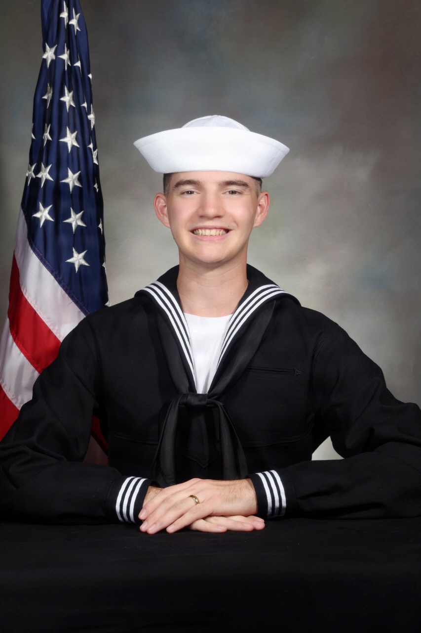 A formal military portrait of a young U.S. Navy enlisted sailor in dress blue uniform (Dress Blues), seated at a black-draped table with hands clasped. An American flag is visible in the background to the left. The subject is smiling and wearing the distinctive white Dixie cup cover.