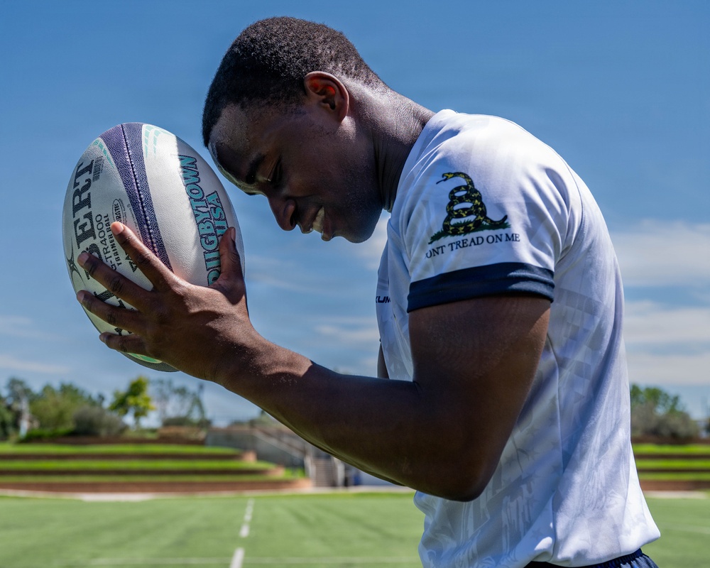 A young man is holding a rugby ball close to his face on what appears to be an athletic field under a clear blue sky. He is wearing a white t-shirt featuring the Gadsden Flag ('Don't Tread on Me') design with a coiled snake on the sleeve. The photo is taken from a low angle, giving a dynamic and close-up perspective.