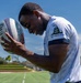 A young man is holding a rugby ball close to his face on what appears to be an athletic field under a clear blue sky. He is wearing a white t-shirt featuring the Gadsden Flag ('Don't Tread on Me') design with a coiled snake on the sleeve. The photo is taken from a low angle, giving a dynamic and close-up perspective.