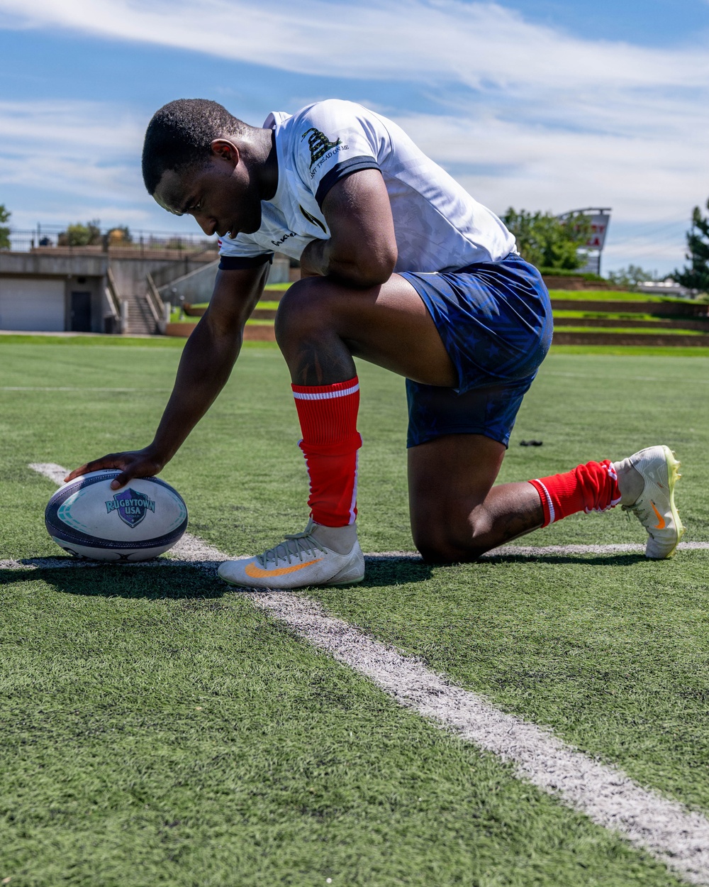 A rugby player is kneeling on an artificial turf field, placing a rugby ball marked 'Rugbytown USA' on a white boundary line. The athlete is wearing a white jersey with a 'Don't Tread on Me' patch, blue shorts, red socks, and Nike cleats. The setting appears to be a stadium or sports complex under a partly cloudy sky.