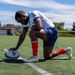 A rugby player is kneeling on an artificial turf field, placing a rugby ball marked 'Rugbytown USA' on a white boundary line. The athlete is wearing a white jersey with a 'Don't Tread on Me' patch, blue shorts, red socks, and Nike cleats. The setting appears to be a stadium or sports complex under a partly cloudy sky.