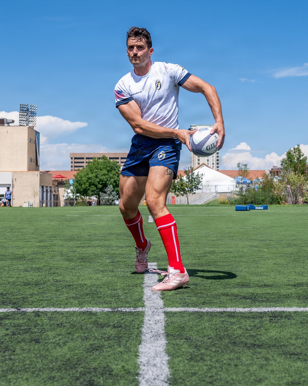 A male rugby player is captured mid-action on an artificial turf field, holding a Gilbert rugby ball in a passing stance. He is wearing a white jersey with an American flag patch and team logo, navy blue shorts, red knee-high socks, and pink/white cleats. The setting appears to be an urban sports facility under bright sunny skies.