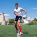 A male rugby player is captured mid-action on an artificial turf field, holding a Gilbert rugby ball in a passing stance. He is wearing a white jersey with an American flag patch and team logo, navy blue shorts, red knee-high socks, and pink/white cleats. The setting appears to be an urban sports facility under bright sunny skies.