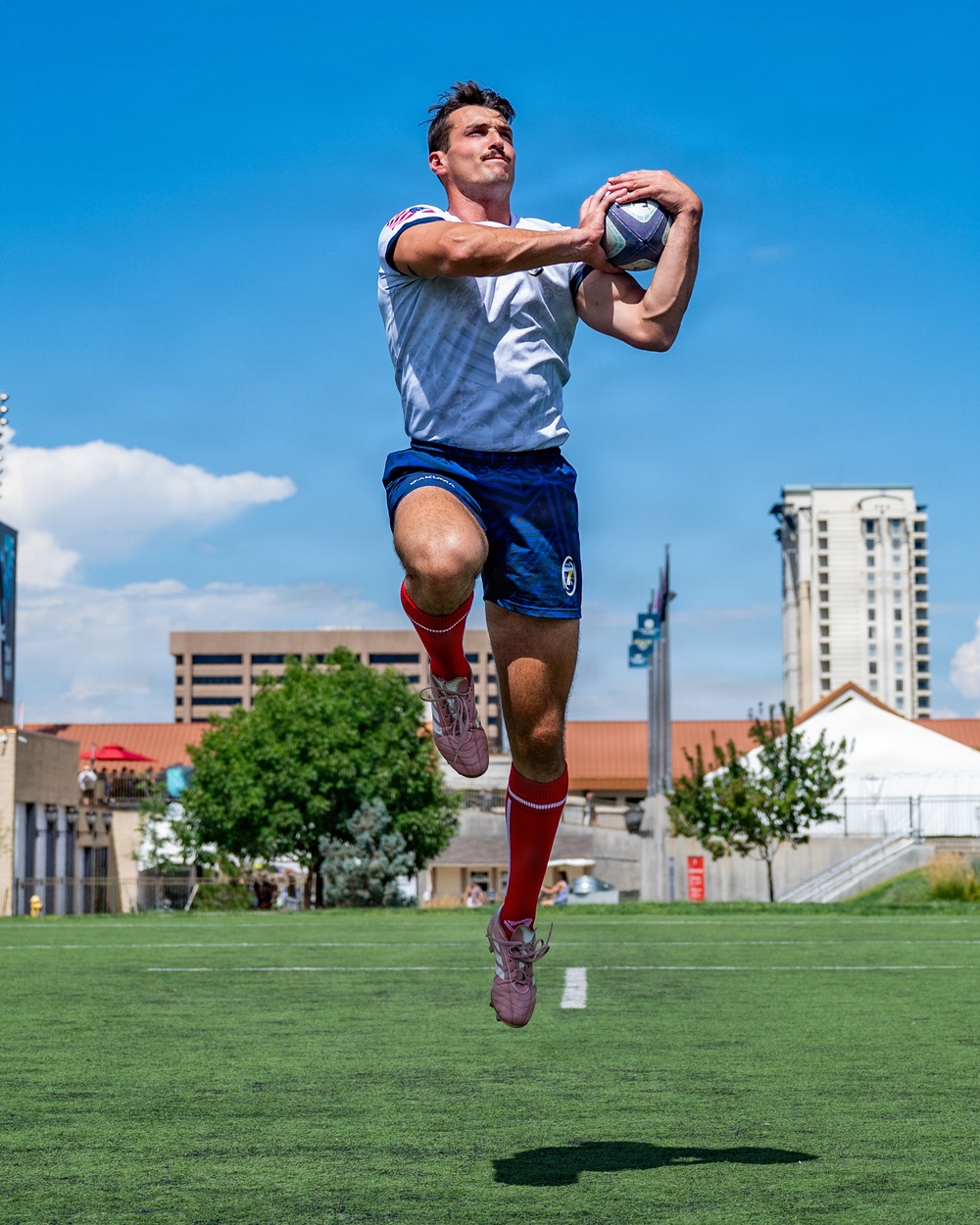 A male rugby player is captured mid-air catching a rugby ball on a green field under a bright blue sky. He is wearing a white jersey, blue shorts, red knee-high socks, and pink/mauve athletic shoes. The background shows an urban setting with multi-story buildings, trees, and spectator areas.