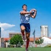 A male rugby player is captured mid-air catching a rugby ball on a green field under a bright blue sky. He is wearing a white jersey, blue shorts, red knee-high socks, and pink/mauve athletic shoes. The background shows an urban setting with multi-story buildings, trees, and spectator areas.