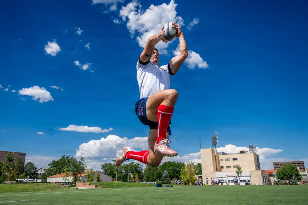 A rugby player is captured mid-air making a high catch of a rugby ball against a bright blue sky with scattered clouds. The athlete is wearing a white jersey, blue shorts, red knee-high socks, and pink/rose gold cleats. The photo is taken from a low angle on a green grass field, with buildings and other players visible in the background.