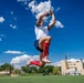 A rugby player is captured mid-air making a high catch of a rugby ball against a bright blue sky with scattered clouds. The athlete is wearing a white jersey, blue shorts, red knee-high socks, and pink/rose gold cleats. The photo is taken from a low angle on a green grass field, with buildings and other players visible in the background.