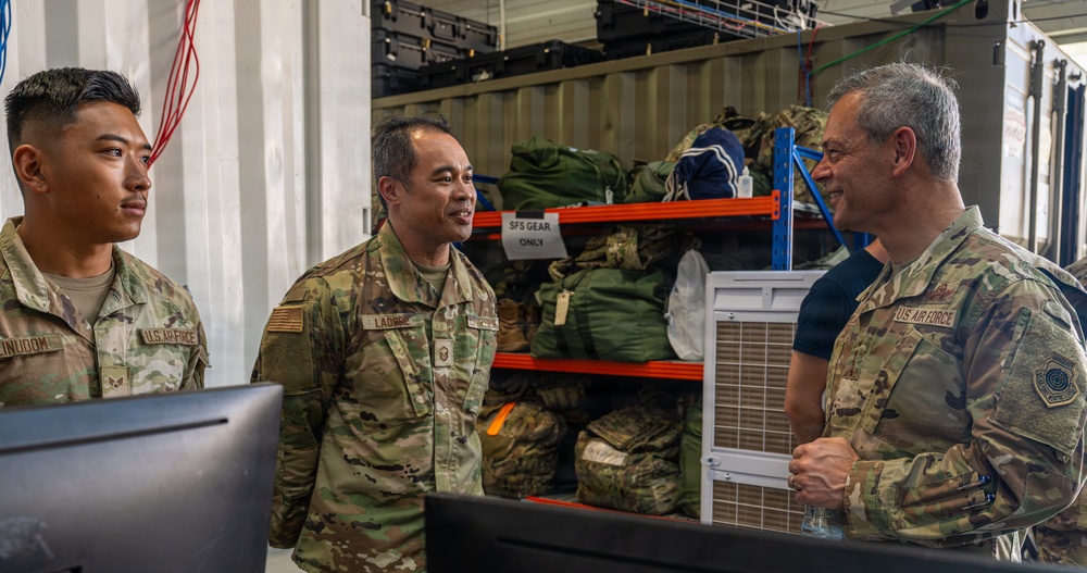 Three U.S. Air Force personnel in OCP (Operational Camouflage Pattern) uniforms are engaged in conversation inside what appears to be a military supply or equipment storage facility. Two of the individuals have visible name tapes, with one reading 'LADRBC' (partially visible) and another partially reading 'INUDOM'. The setting includes shelving units stocked with military gear and a sign reading 'SFS GEAR ONLY'.