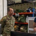 Three U.S. Air Force personnel in OCP (Operational Camouflage Pattern) uniforms are engaged in conversation inside what appears to be a military supply or equipment storage facility. Two of the individuals have visible name tapes, with one reading 'LADRBC' (partially visible) and another partially reading 'INUDOM'. The setting includes shelving units stocked with military gear and a sign reading 'SFS GEAR ONLY'.