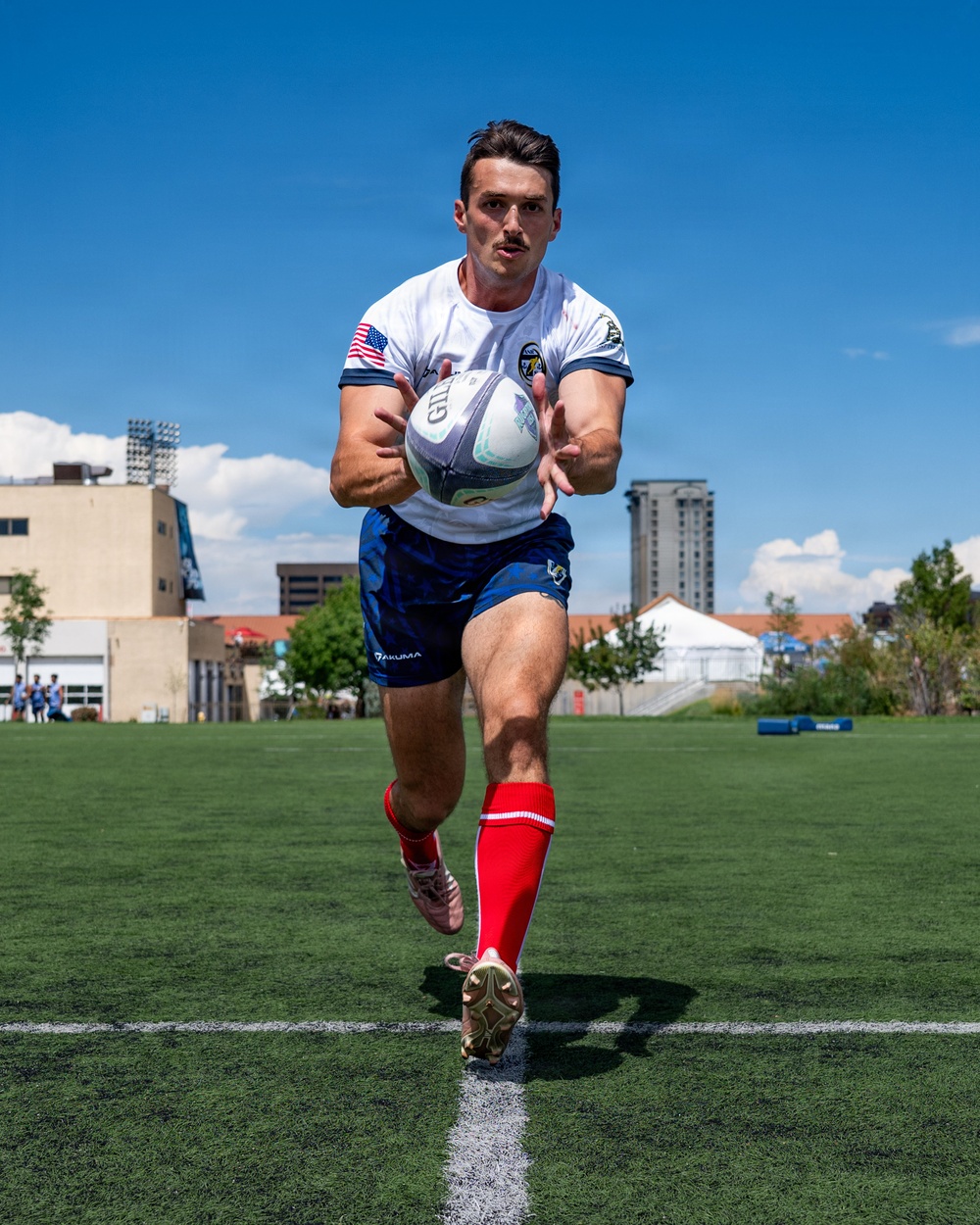 A male rugby player is captured mid-run on a green artificial turf field, carrying a Gilbert rugby ball. He is wearing a white jersey with a US flag patch, blue shorts branded 'AKUMA', red knee-high socks, and gold/pink cleats. The background shows a clear blue sky, urban buildings, and other players in the distance.