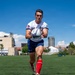 A male rugby player is captured mid-run on a green artificial turf field, carrying a Gilbert rugby ball. He is wearing a white jersey with a US flag patch, blue shorts branded 'AKUMA', red knee-high socks, and gold/pink cleats. The background shows a clear blue sky, urban buildings, and other players in the distance.