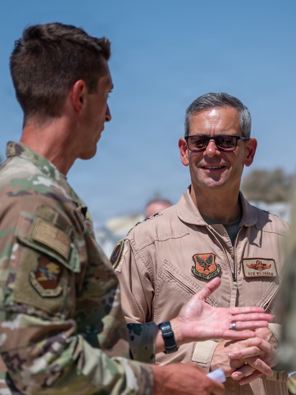 Two U.S. Air Force personnel are engaged in conversation outdoors in what appears to be a deployed or arid environment. The individual on the right is wearing a tan flight suit with a name tape reading 'KEN WILSBACH' and a U.S. Air Force patch, along with general officer rank insignia. The individual on the left is wearing OCP (Operational Camouflage Pattern) uniform.