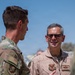 Two U.S. Air Force personnel are engaged in conversation outdoors in what appears to be a deployed or arid environment. The individual on the right is wearing a tan flight suit with a name tape reading 'KEN WILSBACH' and a U.S. Air Force patch, along with general officer rank insignia. The individual on the left is wearing OCP (Operational Camouflage Pattern) uniform.