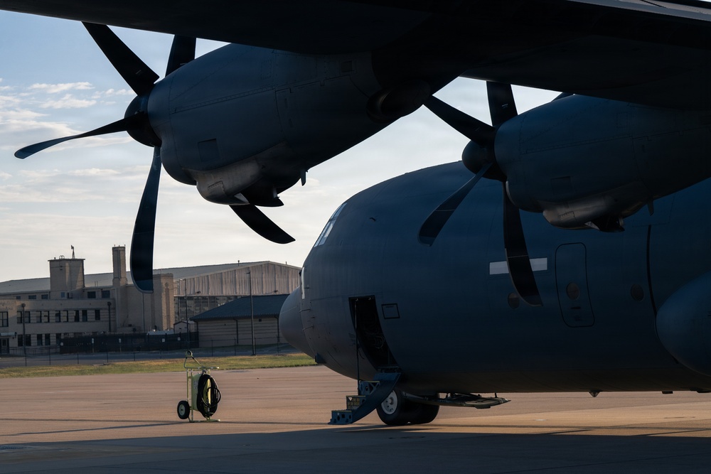A large military turboprop aircraft is parked on a flight line apron, photographed from a low angle beneath the wing. The image captures two visible propeller engines and the fuselage in dark/shadow tones against a partly cloudy sky. Military or airbase infrastructure buildings are visible in the background.