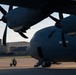 A large military turboprop aircraft is parked on a flight line apron, photographed from a low angle beneath the wing. The image captures two visible propeller engines and the fuselage in dark/shadow tones against a partly cloudy sky. Military or airbase infrastructure buildings are visible in the background.