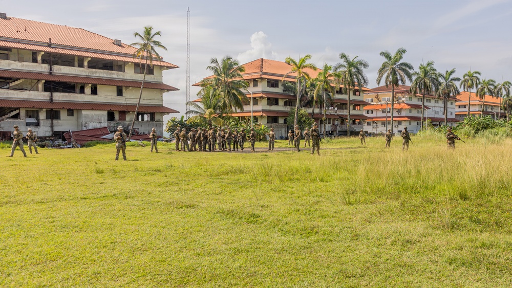 A group of uniformed military or security personnel are assembled on a large grassy field surrounded by multi-story buildings with orange/red tiled roofs and palm trees. The personnel appear to be conducting a formation, briefing, or training exercise in what appears to be a tropical or subtropical location. Some buildings in the background show signs of deterioration or damage.