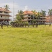 A group of uniformed military or security personnel are assembled on a large grassy field surrounded by multi-story buildings with orange/red tiled roofs and palm trees. The personnel appear to be conducting a formation, briefing, or training exercise in what appears to be a tropical or subtropical location. Some buildings in the background show signs of deterioration or damage.