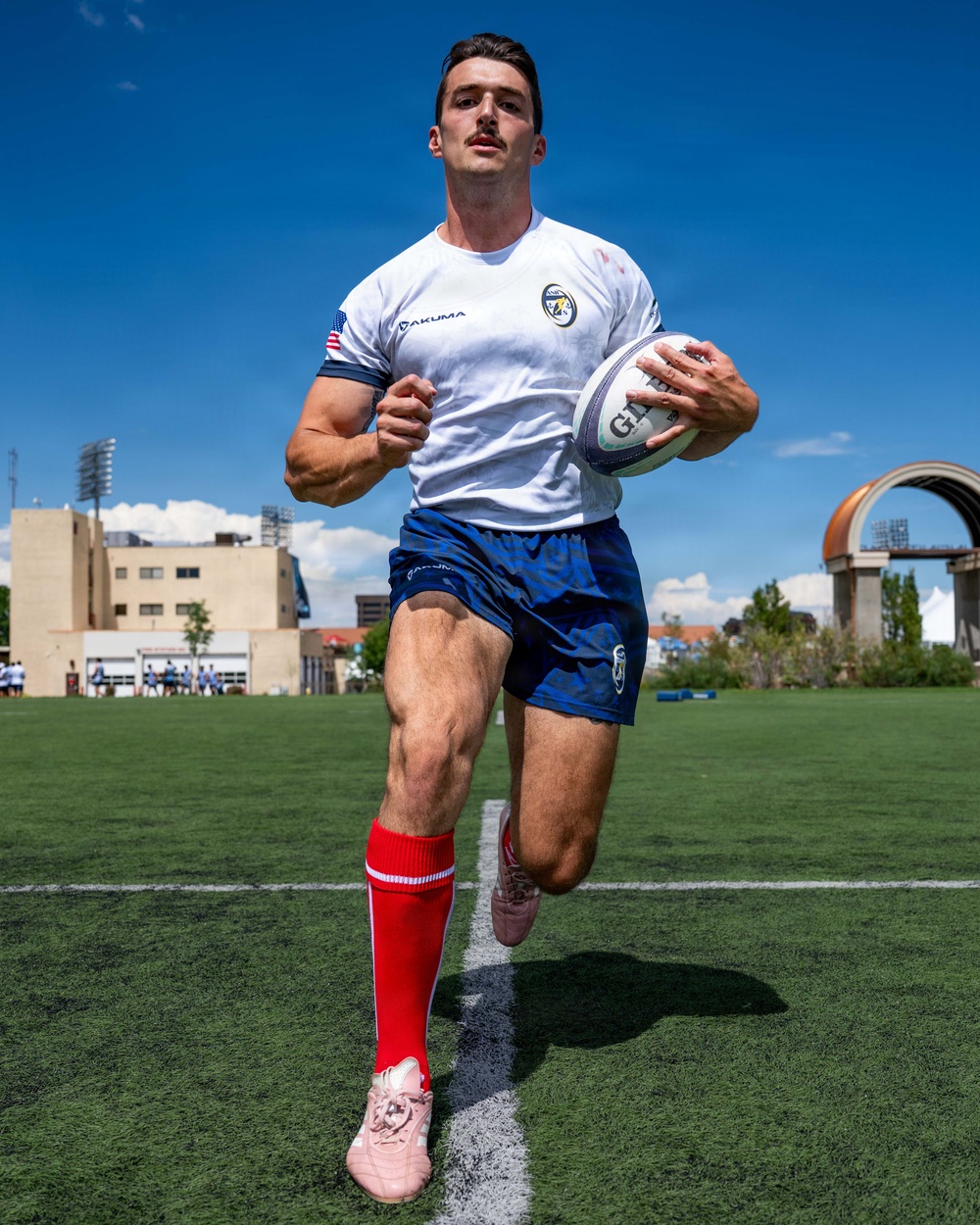 A male rugby player is running with a rugby ball on an artificial turf field under a bright blue sky. He is wearing a white Yakuma jersey with a US flag patch, navy blue shorts, red knee-high socks, and pink cleats. The setting appears to be a competitive rugby venue with stadium lights and urban buildings visible in the background.