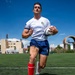 A male rugby player is running with a rugby ball on an artificial turf field under a bright blue sky. He is wearing a white Yakuma jersey with a US flag patch, navy blue shorts, red knee-high socks, and pink cleats. The setting appears to be a competitive rugby venue with stadium lights and urban buildings visible in the background.