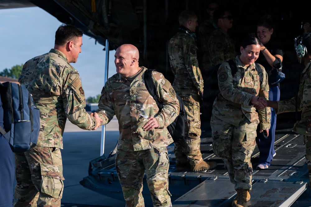 Military personnel in OCP (Operational Camouflage Pattern) uniforms are greeting each other on an airfield tarmac near the rear ramp of a large military transport aircraft. The scene appears to depict a homecoming or arrival event, with handshakes being exchanged between those deplaning and those receiving them. Both male and female service members are present, along with at least one civilian.