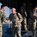 Military personnel in OCP (Operational Camouflage Pattern) uniforms are greeting each other on an airfield tarmac near the rear ramp of a large military transport aircraft. The scene appears to depict a homecoming or arrival event, with handshakes being exchanged between those deplaning and those receiving them. Both male and female service members are present, along with at least one civilian.