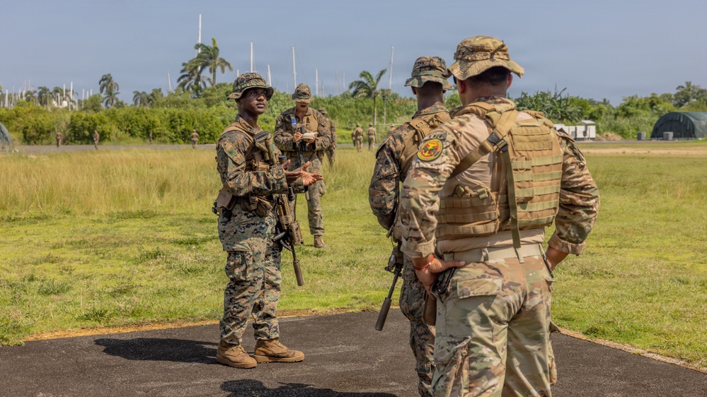 Military personnel in different camouflage uniforms are gathered on a paved surface in a tropical outdoor setting, appearing to be engaged in a briefing or coordination discussion. At least two distinct uniform patterns are visible — MARPAT (Marine pattern) and MultiCam/OCP — suggesting a joint or multinational training exercise. The setting appears to be a military airfield or training area with tropical vegetation in the background.