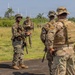 Military personnel in different camouflage uniforms are gathered on a paved surface in a tropical outdoor setting, appearing to be engaged in a briefing or coordination discussion. At least two distinct uniform patterns are visible — MARPAT (Marine pattern) and MultiCam/OCP — suggesting a joint or multinational training exercise. The setting appears to be a military airfield or training area with tropical vegetation in the background.