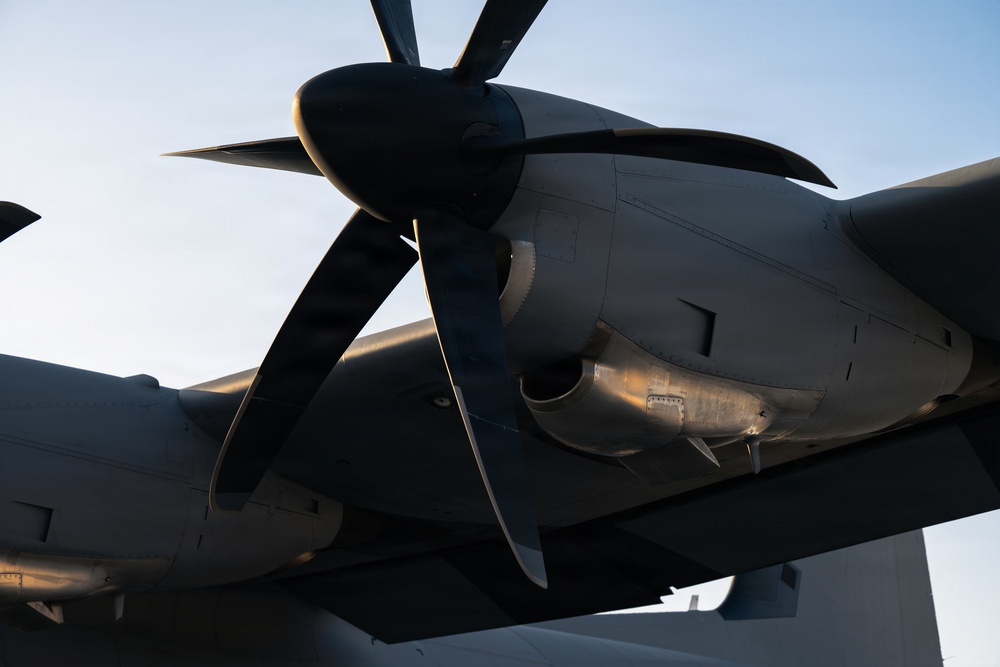 A close-up, low-angle photograph of a large turboprop engine and propeller assembly mounted on an aircraft wing. The image captures the underside of the engine nacelle and multi-blade propeller in dramatic golden-hour lighting. The aircraft appears to be a large military or transport turboprop, stationary on the ground.