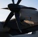 A close-up, low-angle photograph of a large turboprop engine and propeller assembly mounted on an aircraft wing. The image captures the underside of the engine nacelle and multi-blade propeller in dramatic golden-hour lighting. The aircraft appears to be a large military or transport turboprop, stationary on the ground.