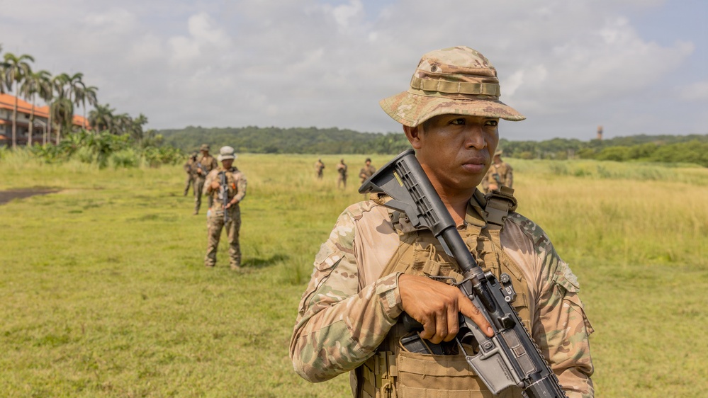 A soldier in multicam camouflage uniform and boonie hat stands in the foreground holding an assault rifle at port arms position. Several other similarly uniformed soldiers are visible in the background on an open grassy field. The setting appears to be a tropical or subtropical environment based on the palm trees and dense vegetation visible.