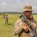 A soldier in multicam camouflage uniform and boonie hat stands in the foreground holding an assault rifle at port arms position. Several other similarly uniformed soldiers are visible in the background on an open grassy field. The setting appears to be a tropical or subtropical environment based on the palm trees and dense vegetation visible.