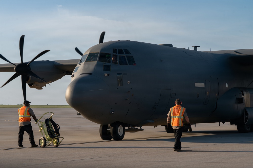 A large military turboprop aircraft, consistent with a C-130 Hercules variant, is parked on a tarmac apron. Two ground crew members wearing orange high-visibility safety vests are approaching the aircraft, with one pulling a wheeled ground support equipment cart. The image is taken during golden hour lighting conditions, casting warm directional light across the scene.