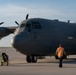 A large military turboprop aircraft, consistent with a C-130 Hercules variant, is parked on a tarmac apron. Two ground crew members wearing orange high-visibility safety vests are approaching the aircraft, with one pulling a wheeled ground support equipment cart. The image is taken during golden hour lighting conditions, casting warm directional light across the scene.