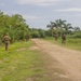 The image shows multiple military or security personnel in camouflage uniforms patrolling along a dirt road in a tropical environment. The soldiers are spread out along both sides of the path, suggesting a security patrol or training exercise. The lush vegetation, palm trees, and humid-looking landscape suggest a tropical or subtropical region.