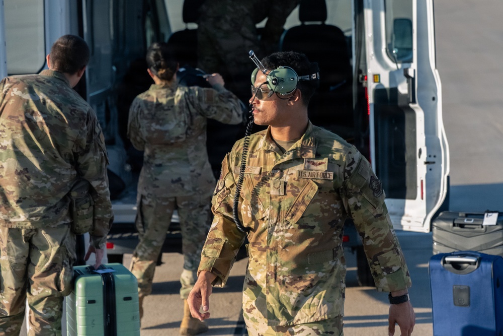 Several U.S. Air Force personnel in OCP (Operational Camouflage Pattern) uniforms are gathered near a white van, apparently loading or unloading equipment and luggage. The foreground airman is notably wearing a flight helmet with visor raised and has a communication cord attached, suggesting aviation-related duties. The scene appears to take place on a flight line or tarmac area.