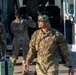 Several U.S. Air Force personnel in OCP (Operational Camouflage Pattern) uniforms are gathered near a white van, apparently loading or unloading equipment and luggage. The foreground airman is notably wearing a flight helmet with visor raised and has a communication cord attached, suggesting aviation-related duties. The scene appears to take place on a flight line or tarmac area.